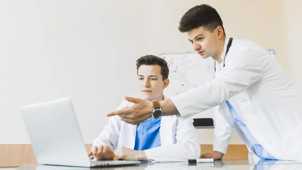 alt="two mbbs students in egypt working on laptop one is sitting infront of the laptop and another one stand beside him. both are wearing white coats